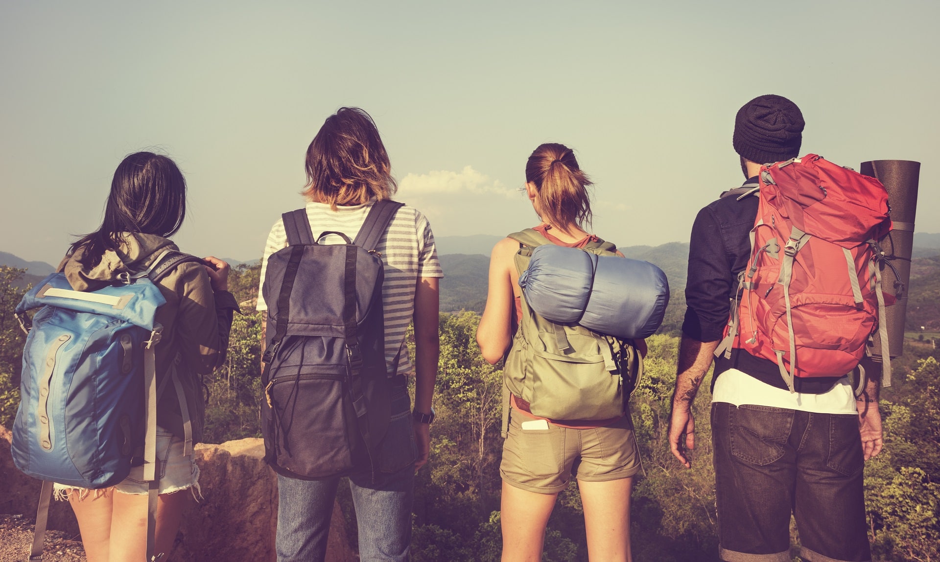 Four hikers with backpacks stand together, gazing at a scenic mountain view under a clear sky.