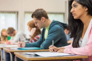 A group of students sits at desks in a classroom, focused on their notebooks and writing materials.