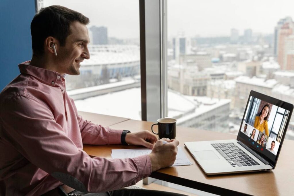 A person in a pink shirt sits at a table with a laptop, headphones, and a coffee cup, attending a virtual meeting with a city view.