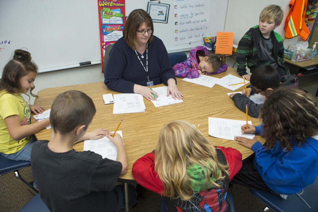 A teacher assists a group of children seated around a table, engaged in a math activity with worksheets and pencils.