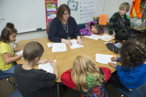 A teacher assists a group of children seated around a table, engaged in a math activity with worksheets and pencils.