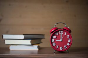 A red vintage alarm clock shows the time as 11:10 beside a stack of books on a wooden table against a blurred background.