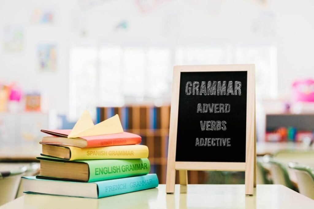 Colorful grammar books titled in various languages stacked beside a chalkboard displaying Grammar, Adverb, Verbs, and Adjective.