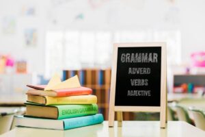 Colorful grammar books titled in various languages stacked beside a chalkboard displaying Grammar, Adverb, Verbs, and Adjective.