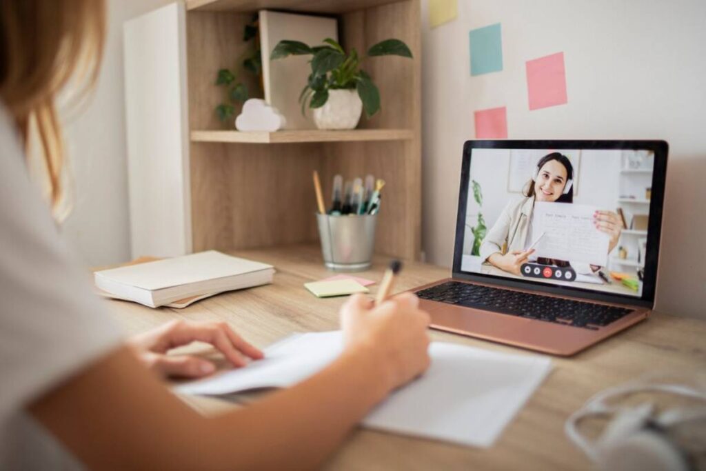 A person is writing notes while attending a video call on a laptop, with a notebook and stationery visible on the desk.