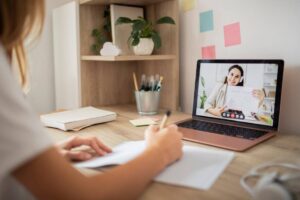 A person is writing notes while attending a video call on a laptop, with a notebook and stationery visible on the desk.