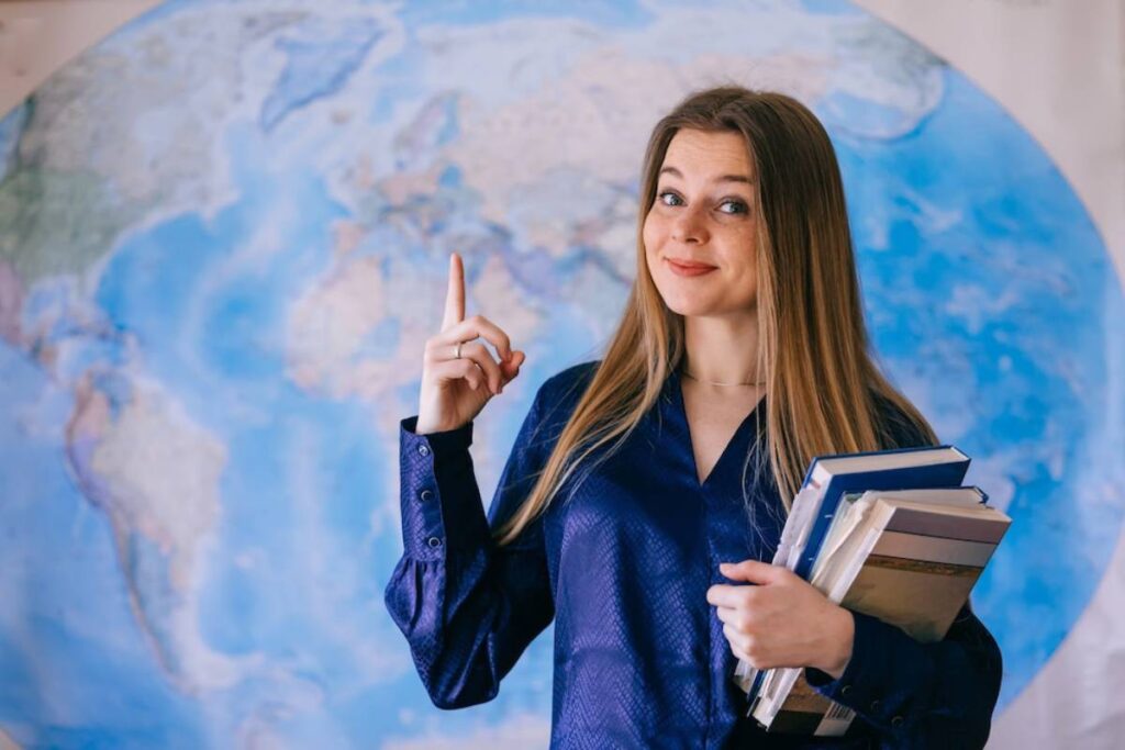 A woman in a blue blouse holds books and gestures upwards, standing in front of a large world map.