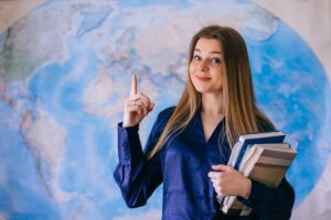 A woman in a blue blouse holds books and gestures upwards, standing in front of a large world map.