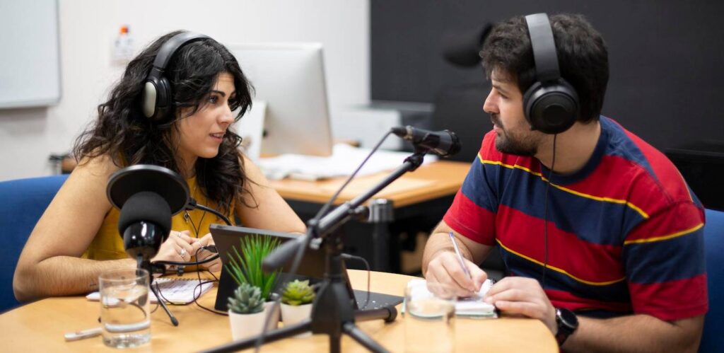 A woman and a man wearing headphones engage in a podcast discussion, seated at a table with a microphone and notes.