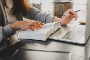 A person in a grey sweater studies from an open book while typing on a laptop, focused on their work in a cozy indoor setting.