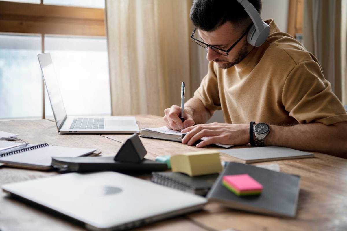 A person wearing headphones writes notes at a wooden desk with a laptop, notebooks, and sticky notes scattered around.