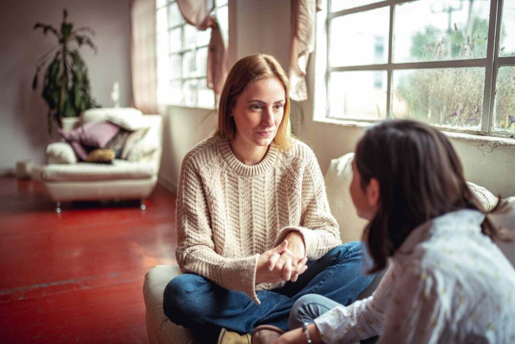 Two women engaged in a deep conversation while sitting on a cushion in a cozy, sunlit room with large windows and a plant in the background.