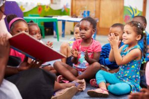 A group of children sits on the floor, attentively clapping hands while engaged in a storytelling session with an adult.