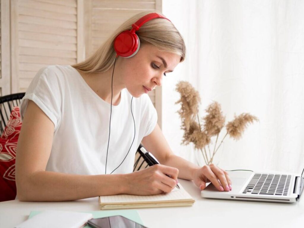A person wearing red headphones writes in a notebook while using a laptop at a bright, cozy workspace with plants in the background.