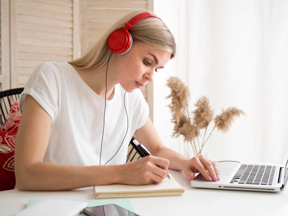 A person wearing red headphones writes in a notebook while using a laptop at a bright, cozy workspace with plants in the background.