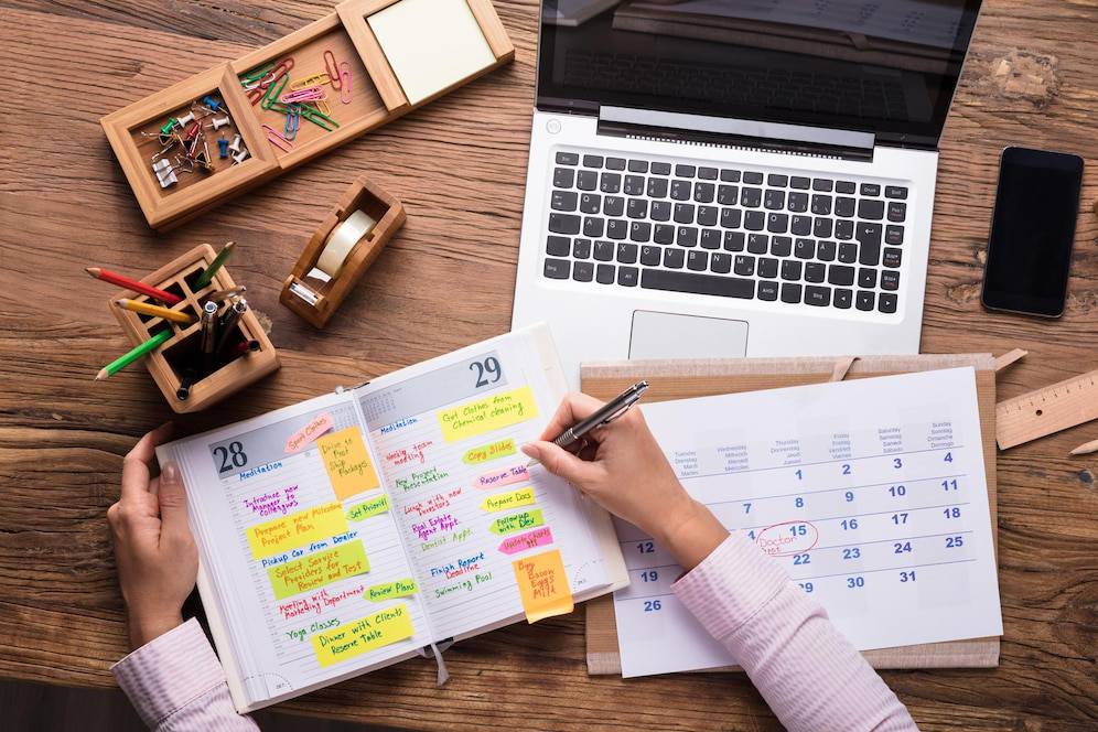 A person writing in a planner filled with colorful notes, surrounded by a laptop, calendar, and stationery on a wooden desk.