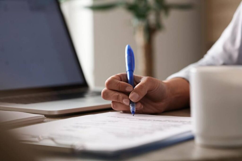A close-up of a hand holding a blue pen, writing notes on paper, with a laptop and a white mug in the background.