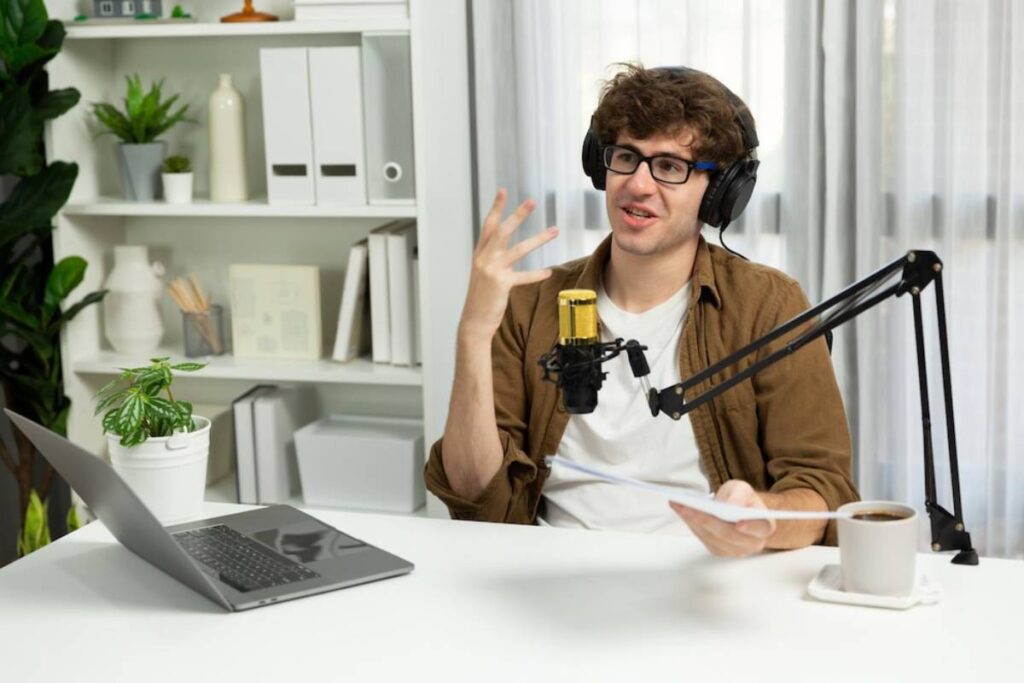 A person with headphones gestures while speaking into a microphone at a desk featuring a laptop, a coffee cup, and green plants.