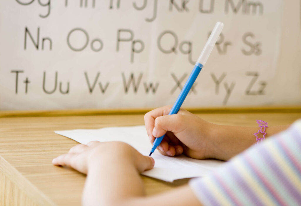 A child writes on paper with a blue pen at a wooden desk, with an alphabet chart displayed in the background.