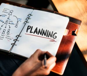A hand holding a pen above an open notebook, prominently featuring the word PLANNING and illustrated diagrams for organization.