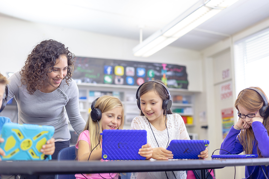 A teacher assists children wearing headphones, engaged with tablets in a colorful classroom setting.