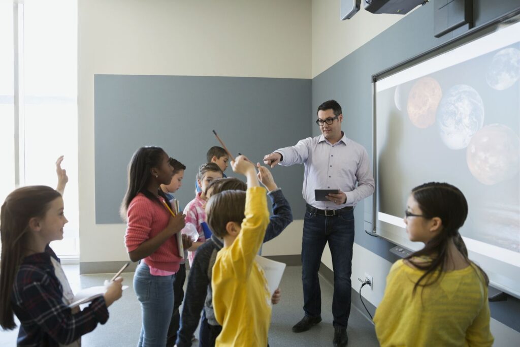 A teacher engages a group of eager students in a classroom, discussing planets displayed on a projector screen.