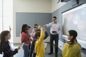 A teacher engages a group of eager students in a classroom, discussing planets displayed on a projector screen.