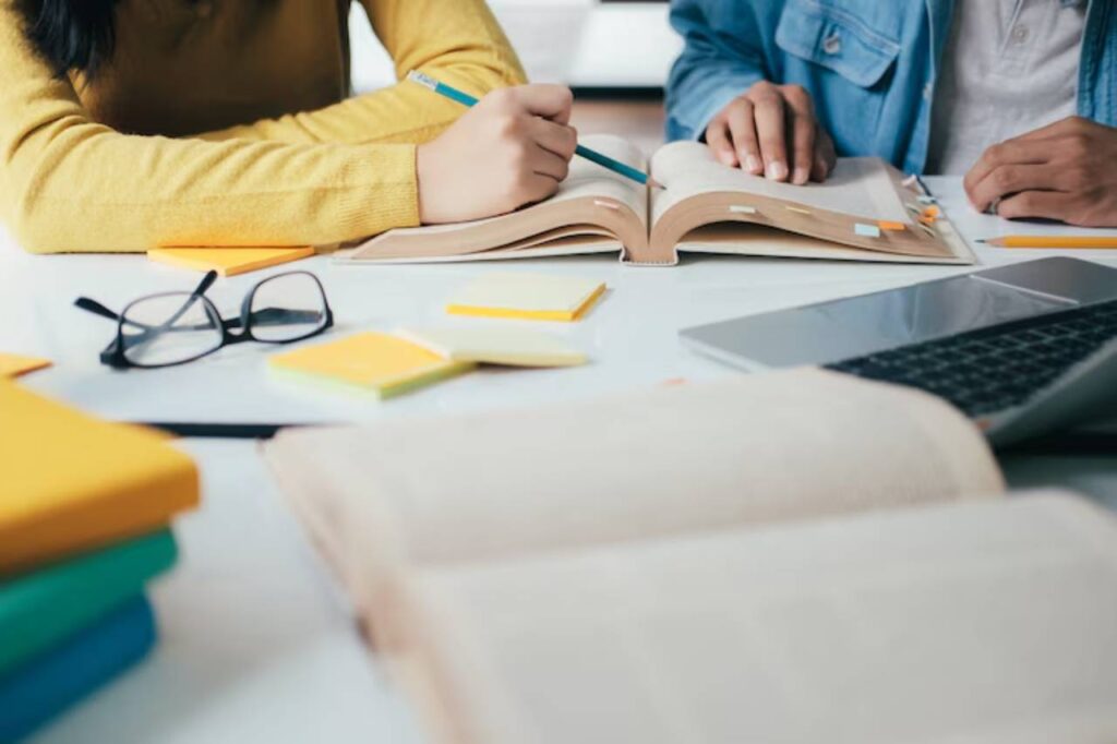 Two people studying together at a table, surrounded by books, sticky notes, eyeglasses, and a laptop. One writes in an open book.