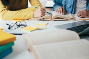 Two people studying together at a table, surrounded by books, sticky notes, eyeglasses, and a laptop. One writes in an open book.