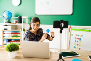 A teacher holds a card displaying a blue ball, with educational materials and a globe visible in a colorful classroom setting.