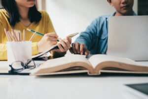 A woman writes in a notebook while a man gestures towards a laptop, with an open book and stationery in the foreground.