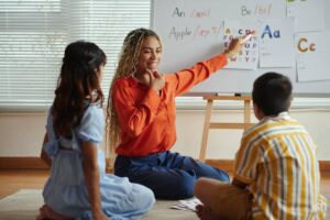 A teacher in an orange shirt engages with two children during a lesson on letters, pointing at an educational chart.