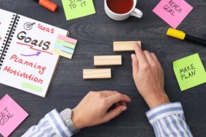 Hands arranging wooden blocks next to a notebook titled Goals and colorful sticky notes, emphasizing planning and motivation.