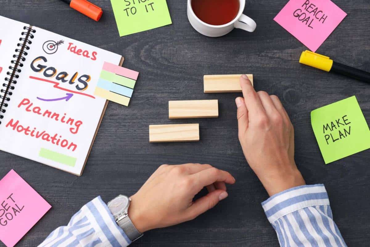 Hands arranging wooden blocks next to a notebook titled Goals and colorful sticky notes, emphasizing planning and motivation.