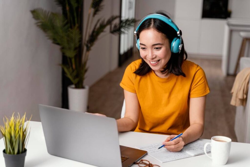 A person in an orange shirt and headphones works at a laptop, taking notes and sipping coffee in a bright, modern workspace.