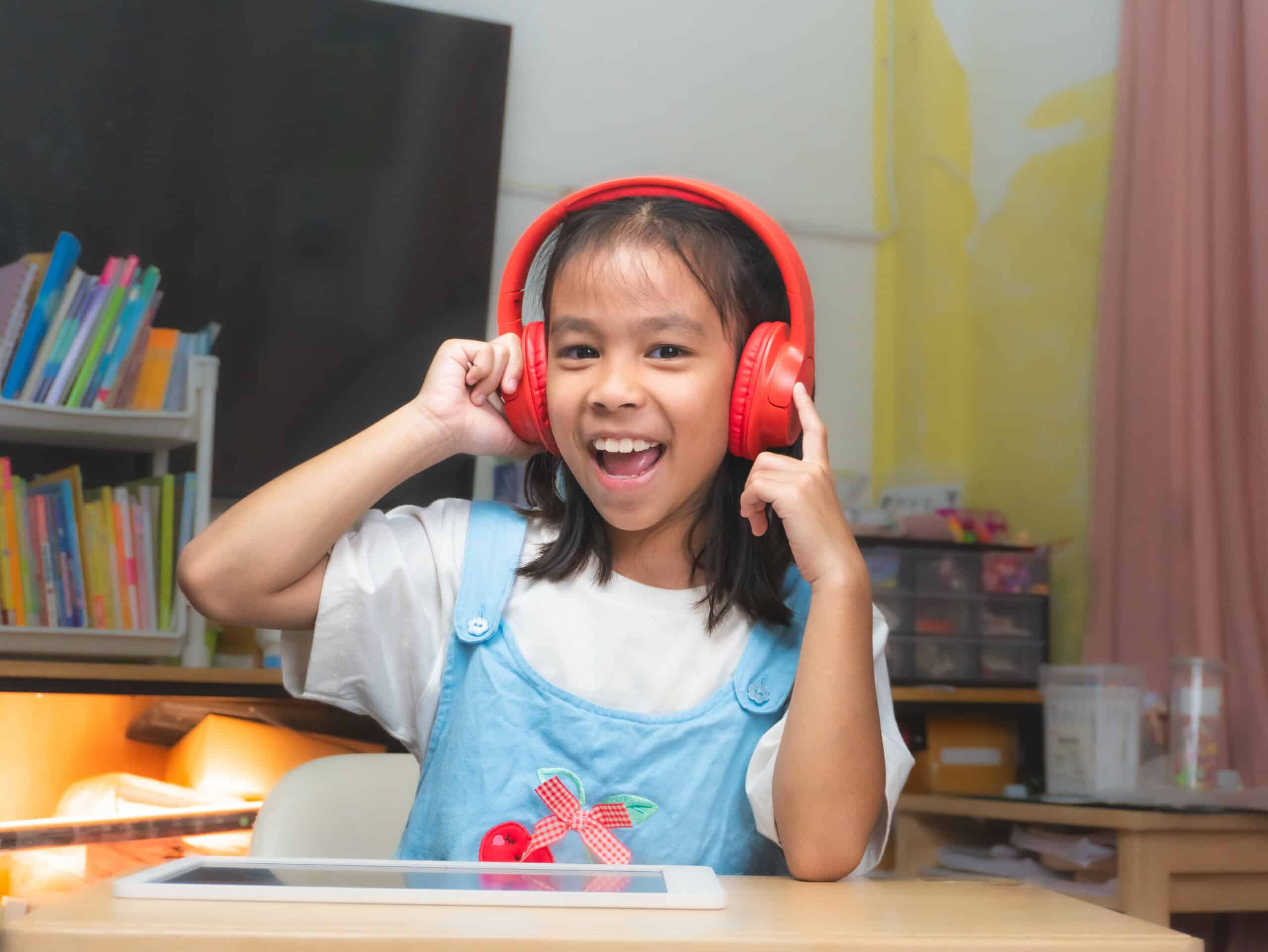 A child wearing red headphones sits at a desk, with a tablet in front and colorful books in a nearby shelf. Bright, playful decor surrounds them.