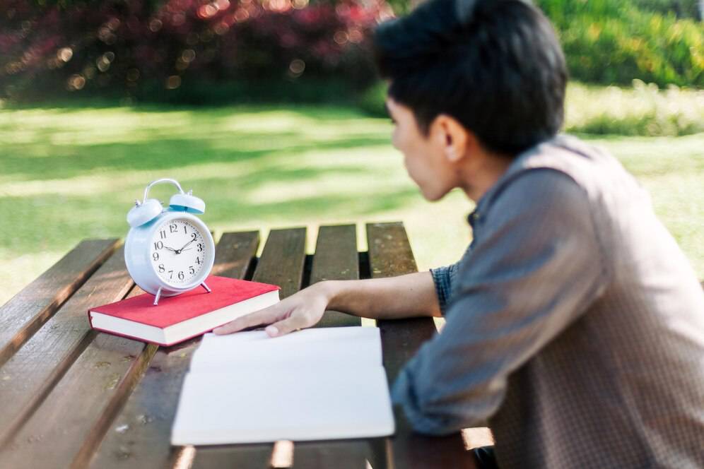 A person sits at a wooden table outdoors, gazing at a vintage blue alarm clock next to a red book and a blank notepad. 