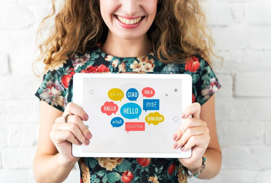 A smiling woman with curly hair holds a tablet displaying various greetings in multiple languages against a white brick wall. 