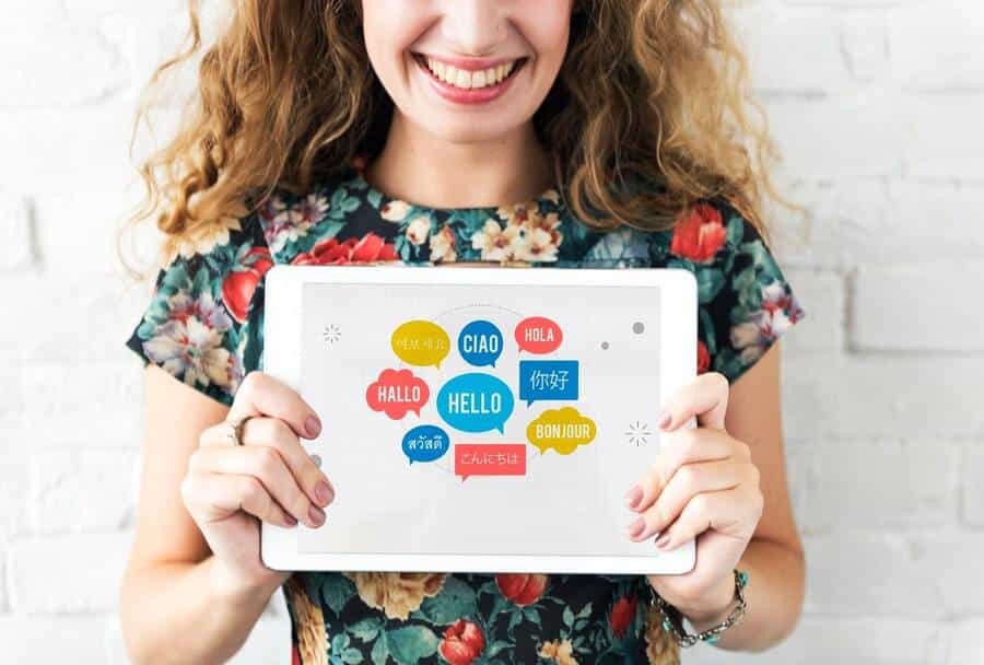 A smiling woman with curly hair holds a tablet displaying various greetings in multiple languages against a white brick wall.