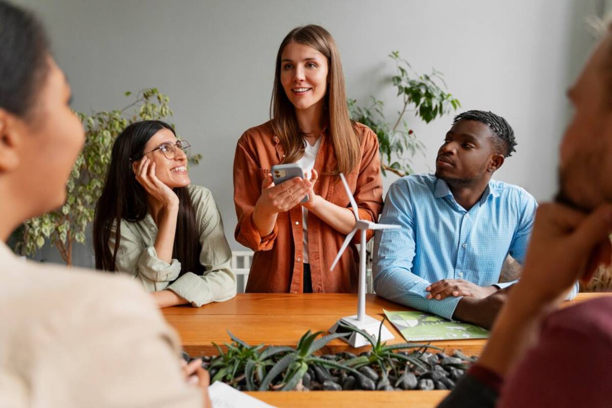  A woman stands at a table discussing ideas, while colleagues listen and a small wind turbine model is visible among plants.