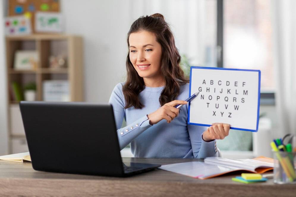  A woman in a blue sweater points to a chart of the English alphabet while sitting at a desk with a laptop and colorful stationary.