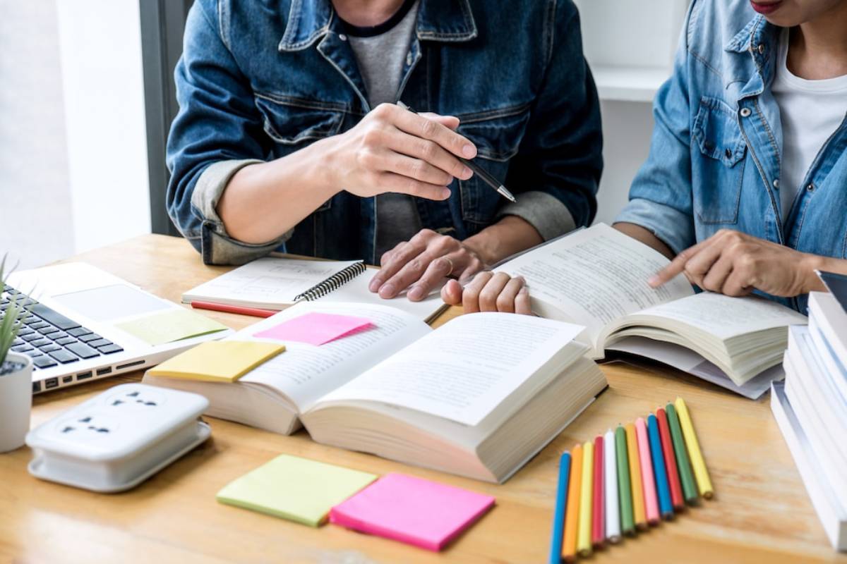  Two people studying together at a table, surrounded by open books, sticky notes, a laptop, and colorful pens.