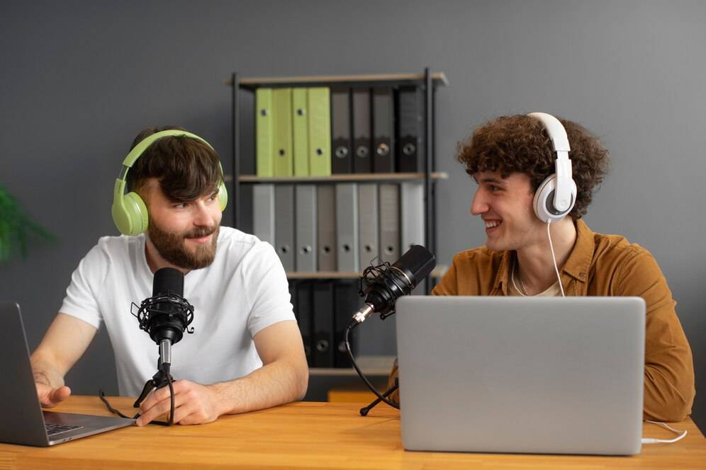  Two podcast hosts with microphones and headphones sit at a wooden table, laptops in front, surrounded by office supplies and folders.