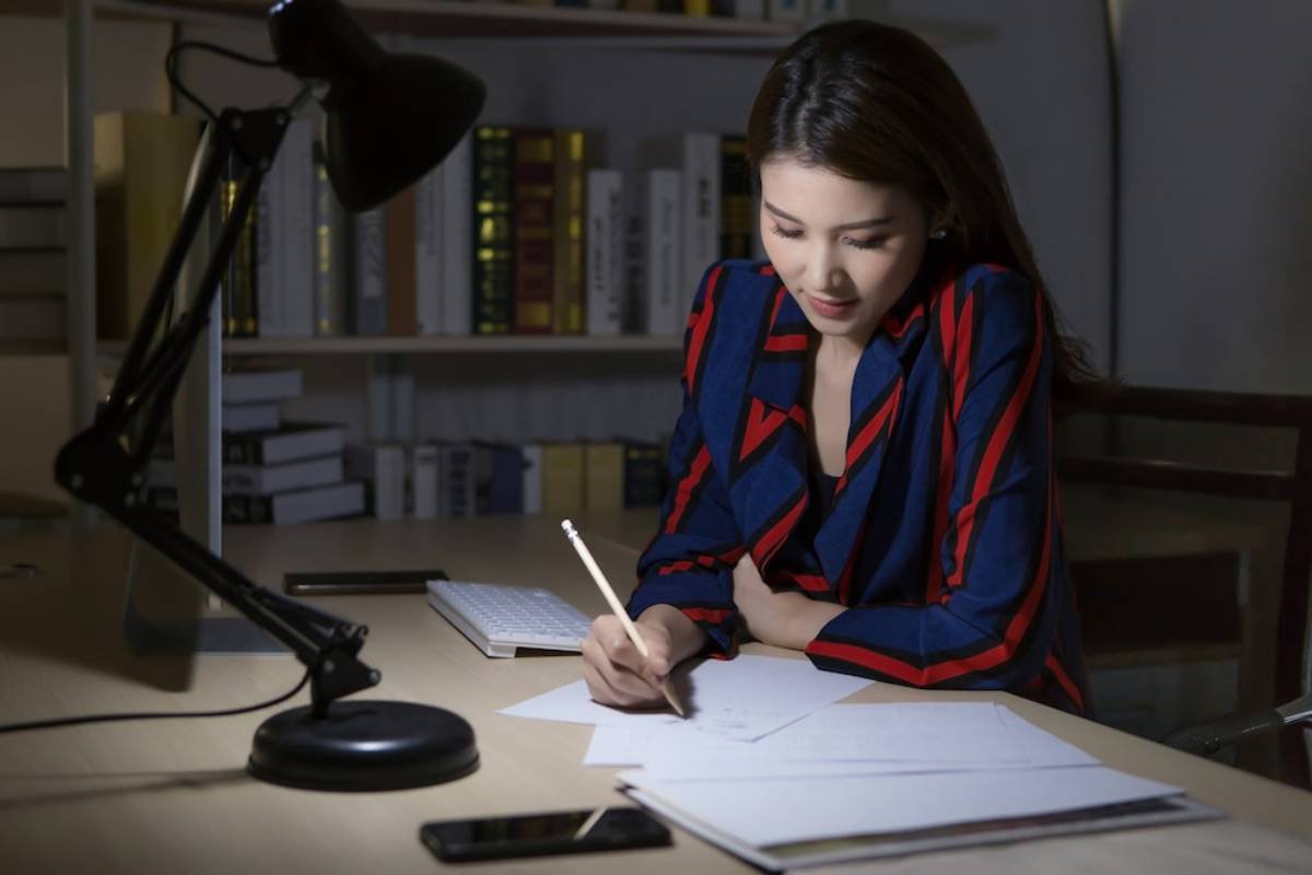 A woman in a blue and red striped blazer sits at a desk, writing on sheets of paper, illuminated by a desk lamp. Books are visible in the background.