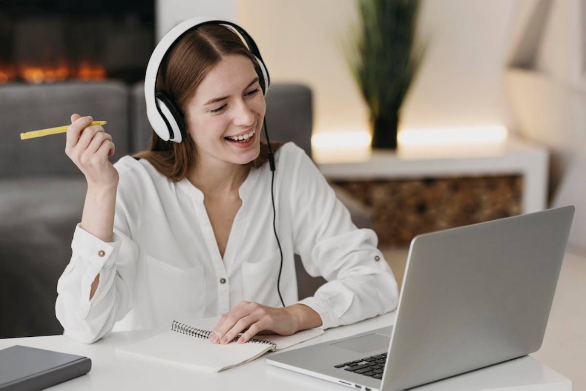 A person wearing headphones sits at a desk, holding a yellow pencil, with a laptop and notepad in a cozy, well-lit room. 