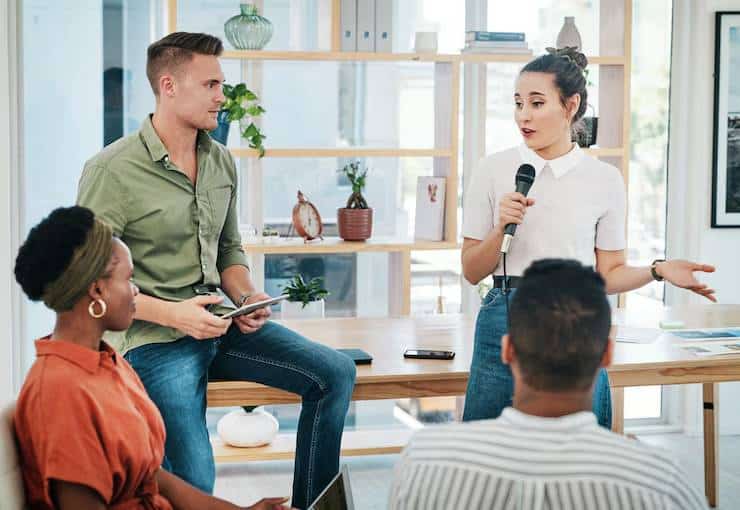 A woman speaks into a microphone while presenting to a group seated around a table in a modern workspace setting.