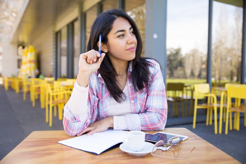 A woman sitting at an outdoor café table, resting her chin on a hand, with a notebook, coffee, and smartphone in front of her. 