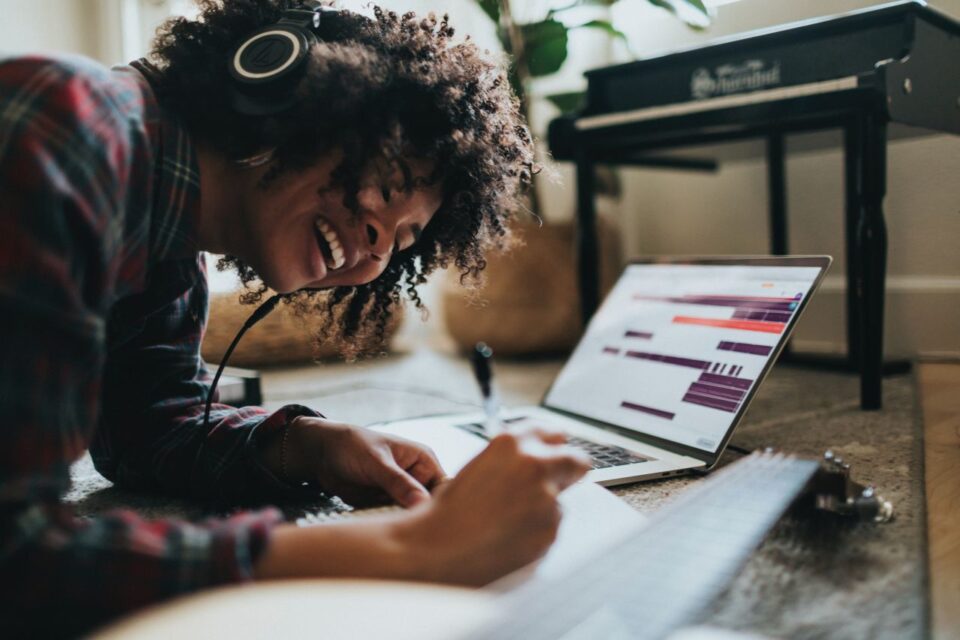  A young woman with curly hair smiles while writing notes in a notebook, headphones on, next to a laptop displaying music software.
