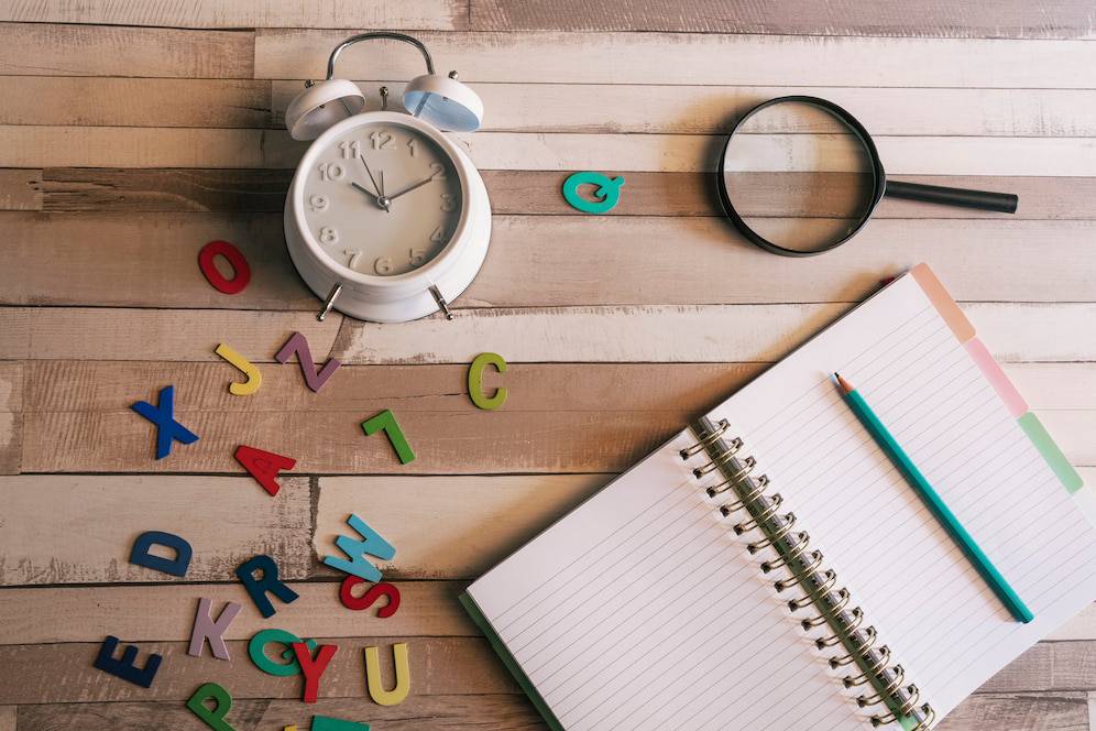 A white alarm clock, colorful letters, a magnifying glass, and a spiral notebook with a turquoise pencil on a wooden surface. 