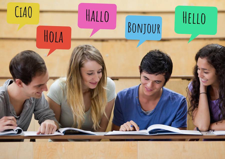 Four students are studying together at a desk, with colorful speech bubbles displaying greetings in various languages. 